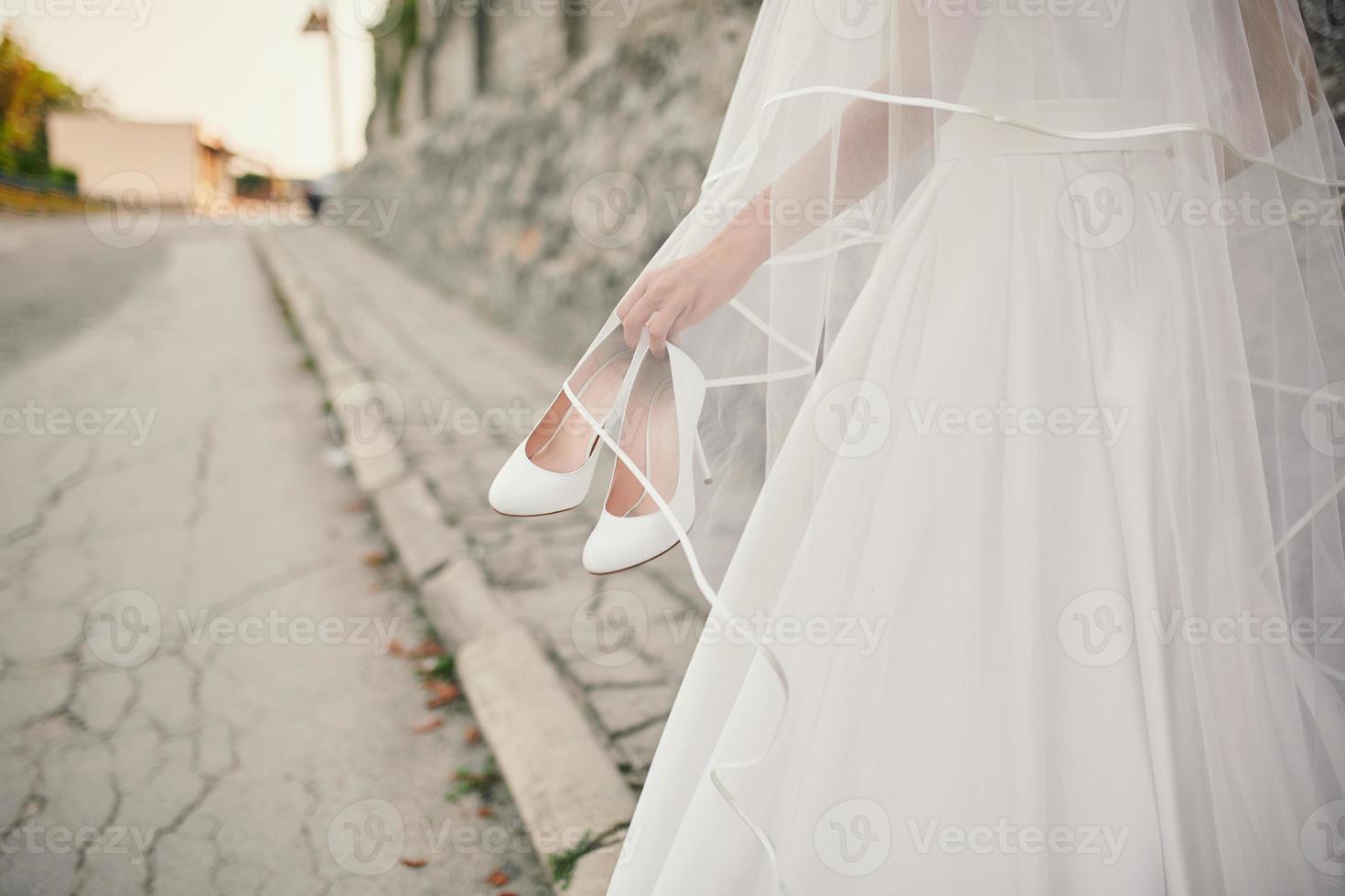 la mariée vêtue d'une robe de mariée et d'un voile marche dans la rue avec des chaussures à la main photo