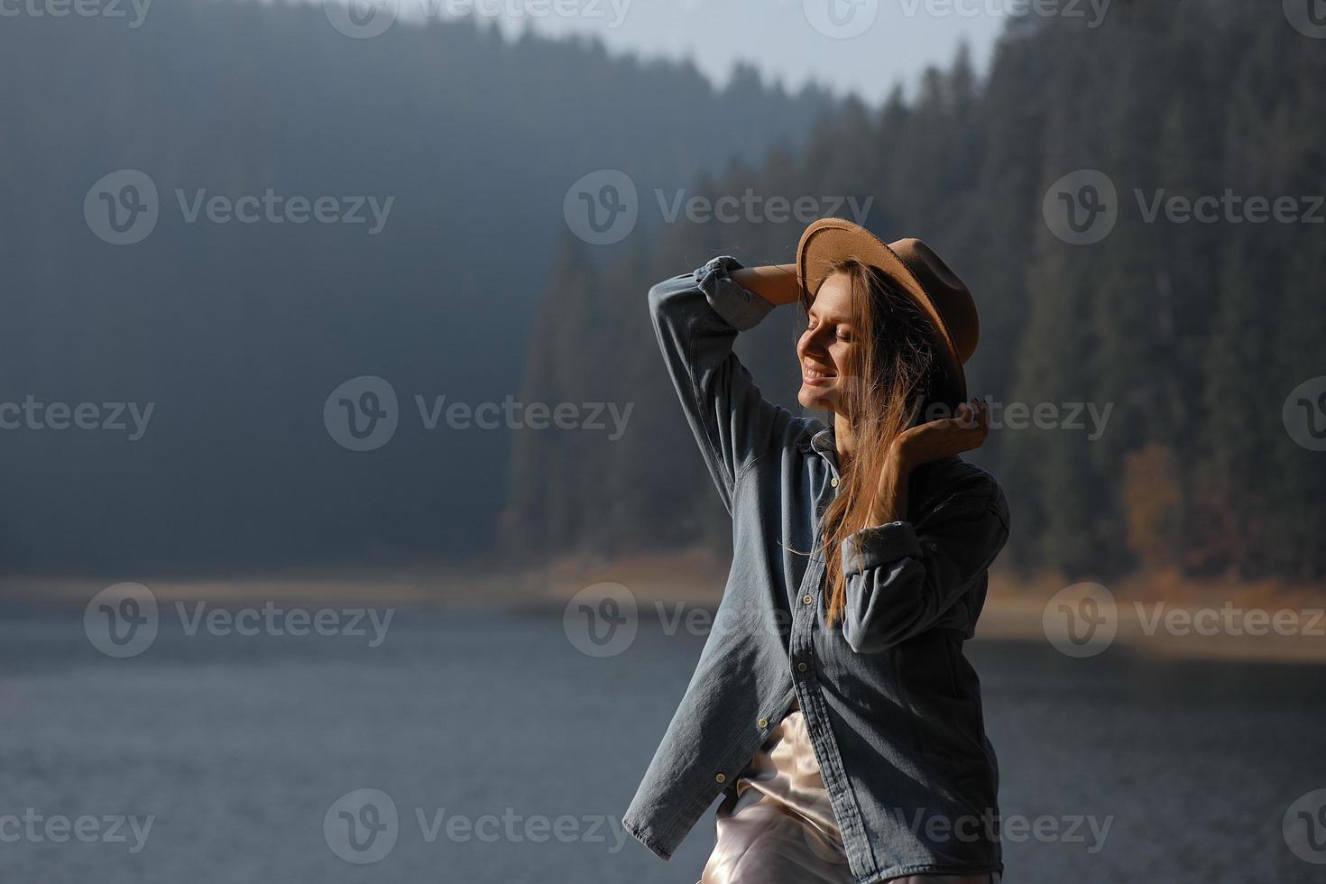 heureuse jeune femme au chapeau bénéficie d'une vue sur le lac dans la forêt. moments de détente. vue d'une fille élégante profite de la fraîcheur en plein air. liberté, personnes, style de vie, voyages et vacances photo