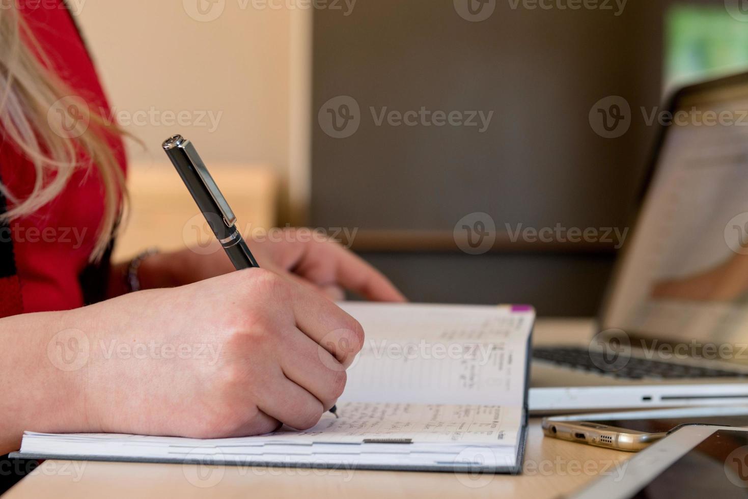 femme blonde assise devant un ordinateur portable et écrivant. il y a un ordinateur portable, une tablette, un téléphone et un cahier sur la table. photo