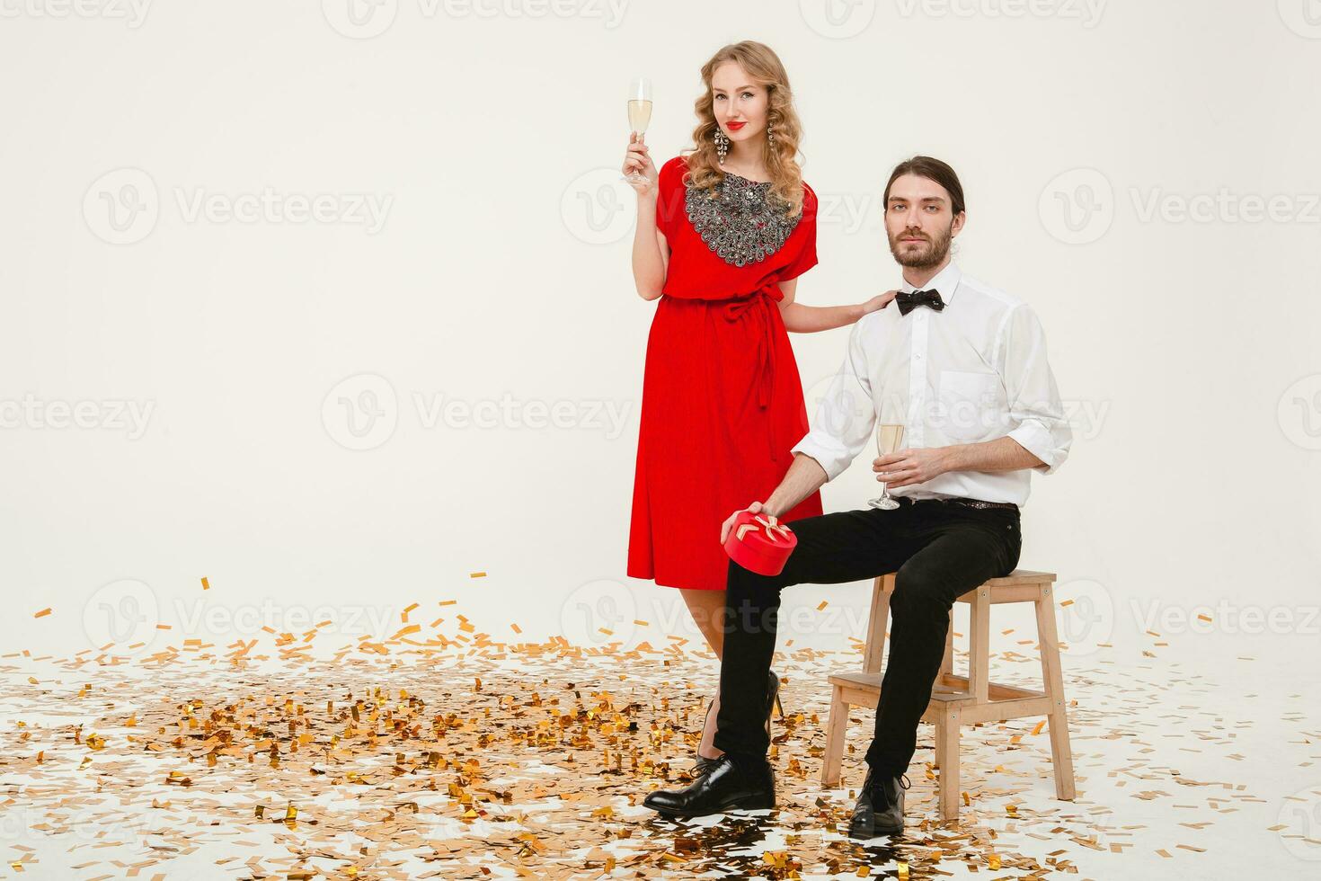 Jeune élégant couple dans l'amour sur blanc Contexte en portant des lunettes et en buvant Champagne, célébrer Nouveau année, portant rouge robe, mode bijoux, arc cravate, content disco faire la fête, ayant amusement photo