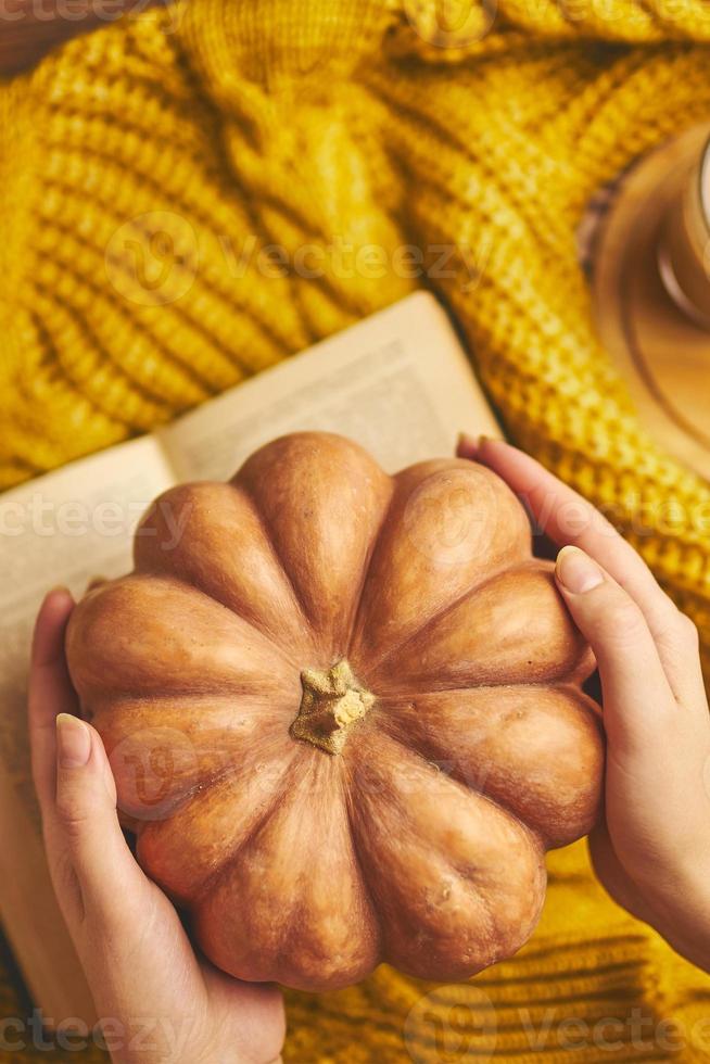 citrouille mûre ronde dans les mains de la femme, livre ouvert et pull tricoté chaud. photo