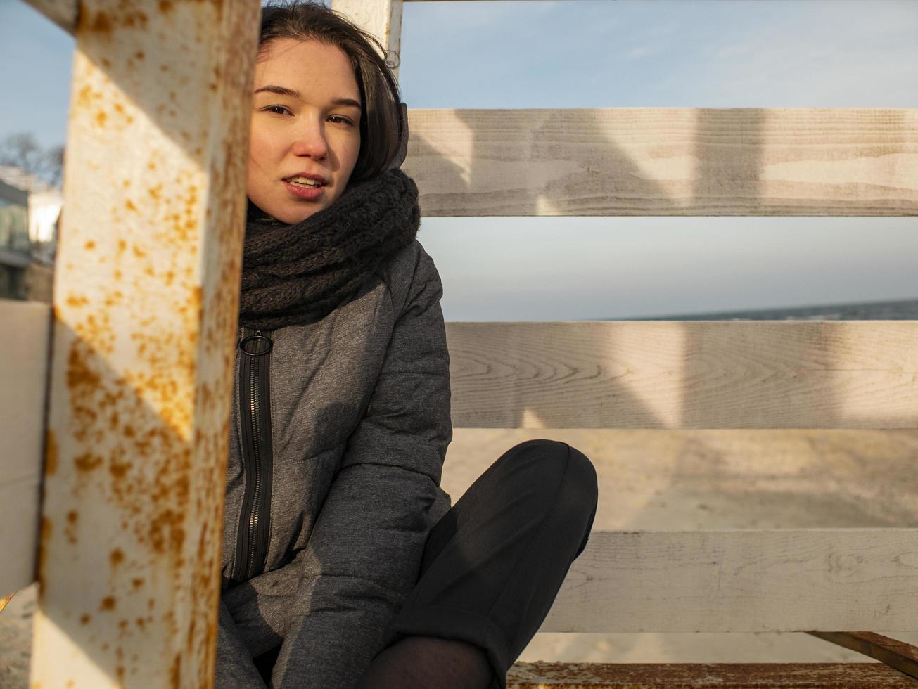 Jolie jeune fille aux cheveux ébouriffés est assise sur un design en bois sur la plage dans un manteau photo