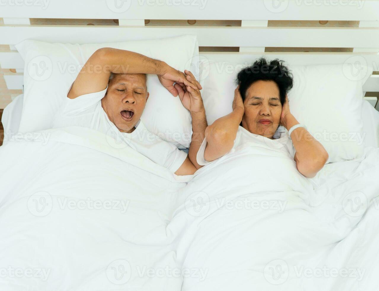 plus âgée asiatique femme pose ouvert sa yeux et couverture oreilles avec sa mains dans lit à côté de sa mari qui ronfle et fait du bruit, matrimonial problèmes. photo