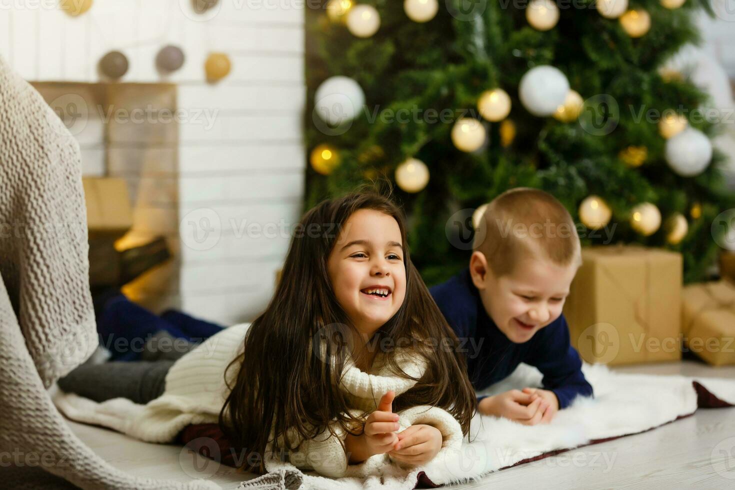 famille sur Noël veille à cheminée. les enfants en dessous de Noël arbre avec cadeau des boites. décoré vivant pièce avec traditionnel Feu lieu. confortable chaud hiver soir à maison. photo