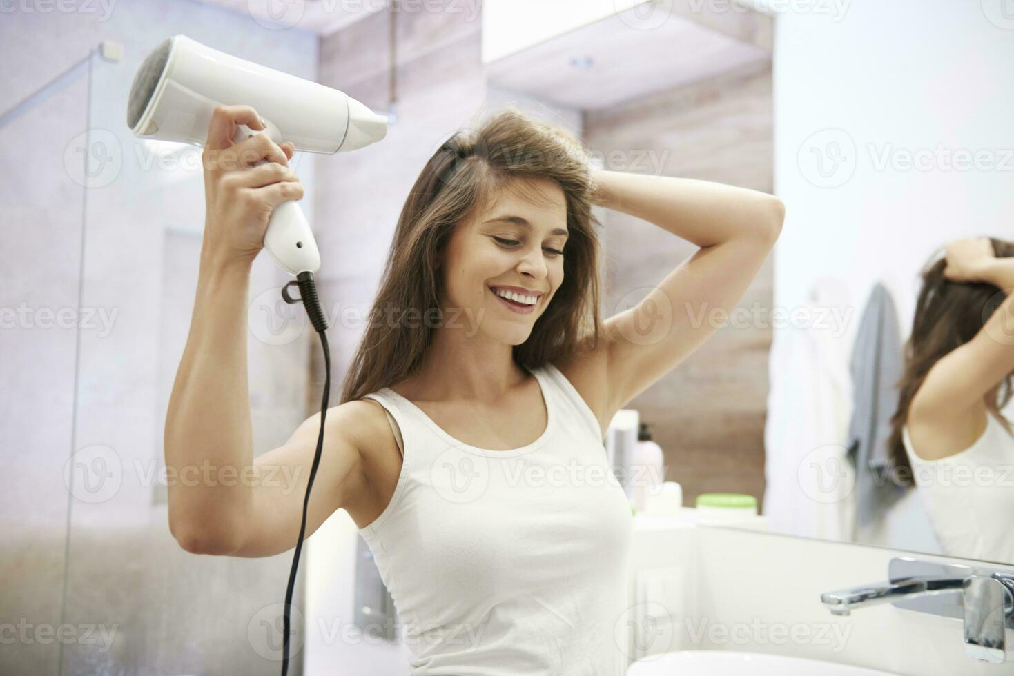 portrait de en riant femme en utilisant cheveux séchoir dans le salle de bains photo