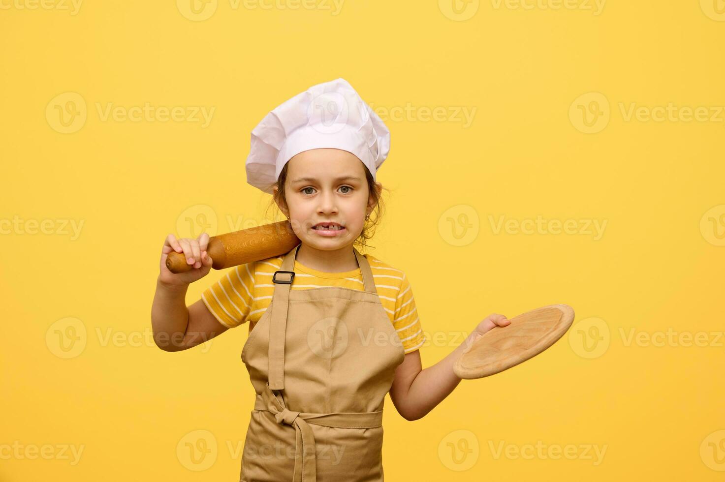 magnifique caucasien peu fille fabrication visages, à la recherche à caméra, en portant roulant épingle et en bois conseil, isolé Jaune photo