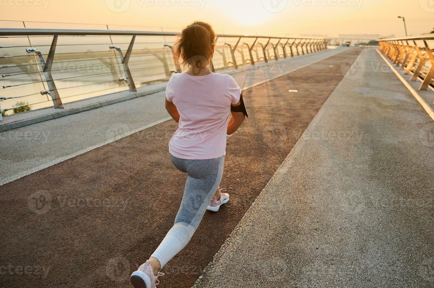 arrière vue de une femelle athlète dans tenue de sport élongation sa jambes muscles, performant fentes, travail en dehors sur une ville pont dans le de bonne heure Matin à lever du soleil. Jeune sportive exercice Extérieur à aube. photo