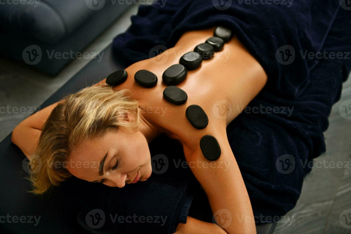 magnifique européen Jeune blond femme avec magnifique visage mensonge sur une massage table avec chaud lave des pierres sur sa retour le long de sa colonne vertébrale recevoir ayurvédique traitement dans une bien-être spa clinique photo