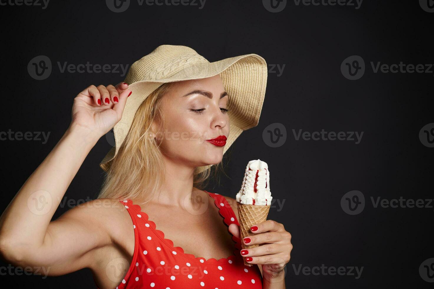 attrayant blond caucasien femme portant paille été chapeau et rouge maillot de bain avec blanc polka des points, en portant la glace crème cône et profiter chaud été journées isolé plus de noir Contexte avec copie un d espace photo
