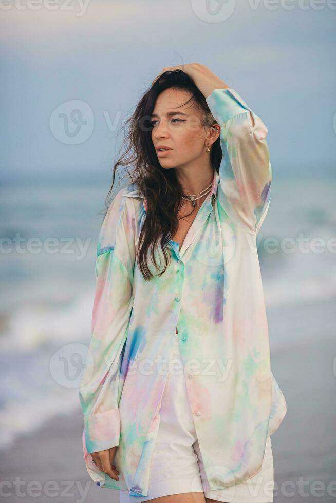 Jeune content femme sur le plage prendre plaisir sa été vacances. femme en marchant le long de le mer dans soir. photo