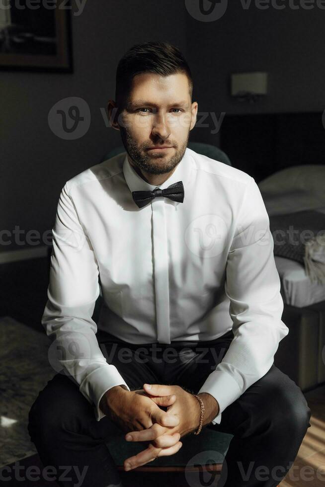 le élégant jeune marié pose à la recherche à le caméra, en train de préparer pour une mariage cérémonie. grand portrait. magnifique intérieur dans le Matin lumière. le du marié Matin photo