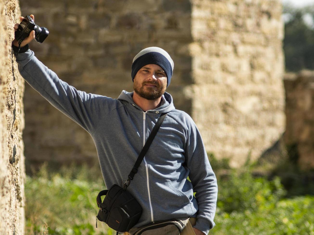 un homme barbu se tient contre un mur dans une forteresse avec un appareil photo