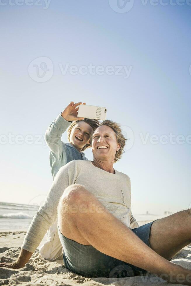 père ayant amusement avec le sien fils sur le plage, prise téléphone intelligent des photos