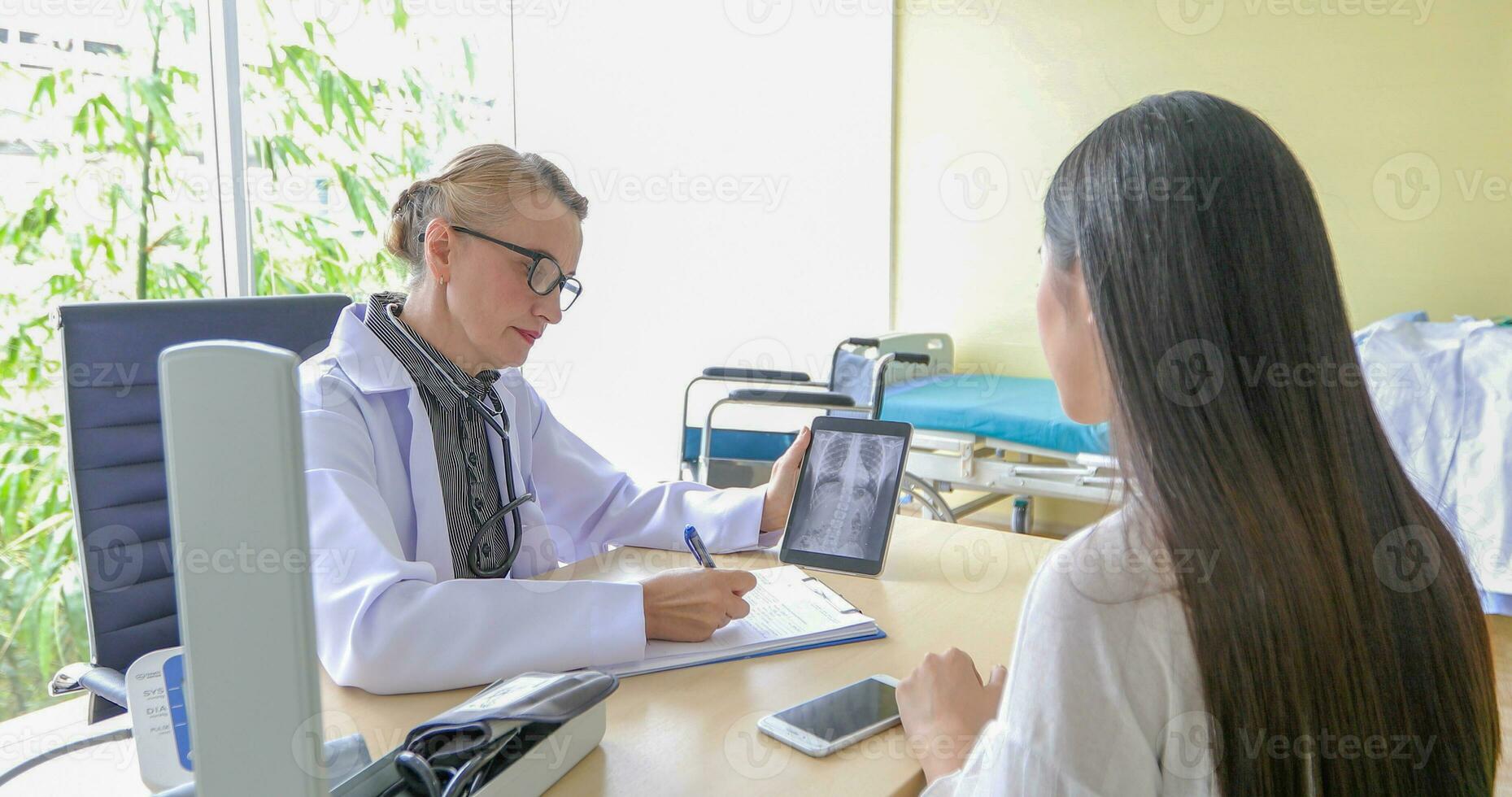 asiatique femme patient séance avec médecin à propos sa maladies et montrant radiographie résultats avec du sang pression et cœur taux la mesure avec stéthoscope photo