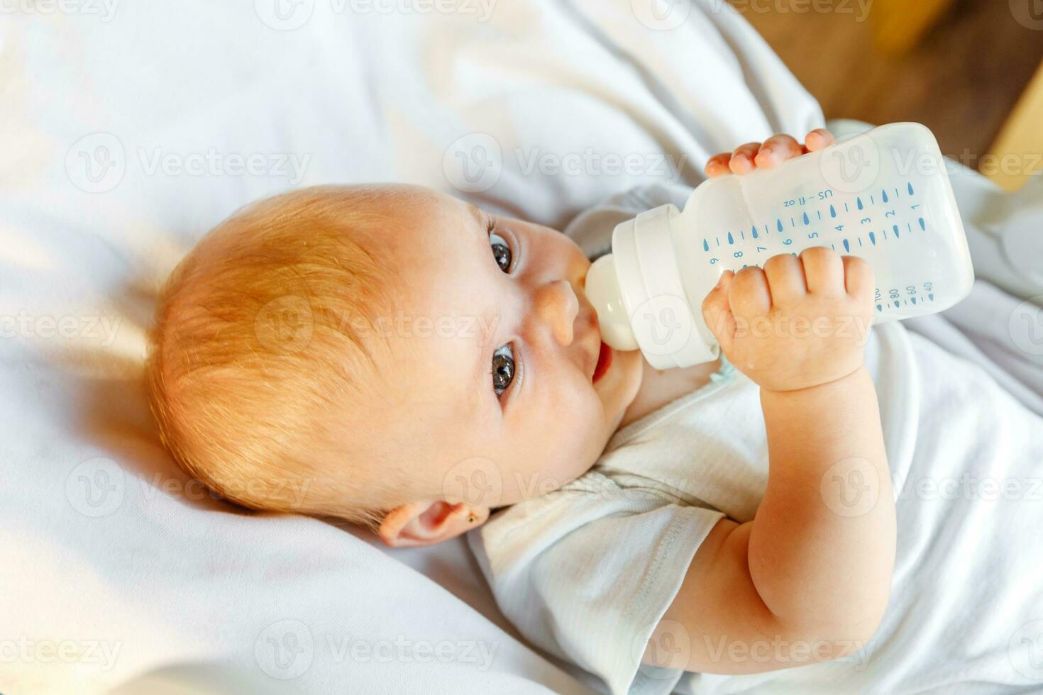 mignonne peu nouveau née fille en buvant Lait de bouteille et à la recherche à caméra sur blanc Contexte. bébé bébé succion en mangeant Lait nutrition mensonge vers le bas sur lit de bébé lit à maison. maternité content enfant concept. photo