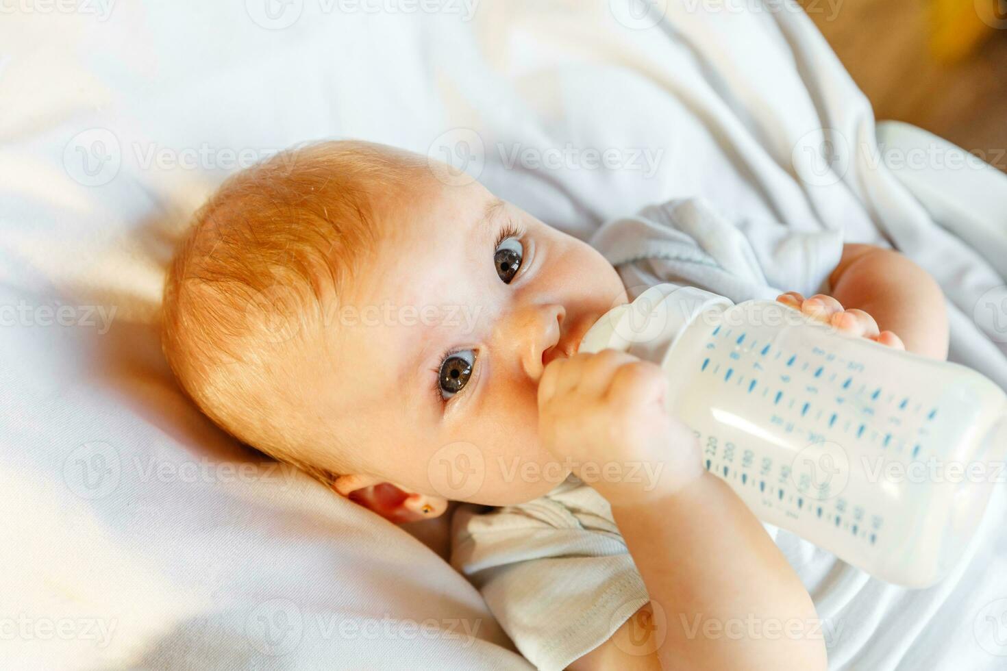 mignonne peu nouveau née fille en buvant Lait de bouteille et à la recherche à caméra sur blanc Contexte. bébé bébé succion en mangeant Lait nutrition mensonge vers le bas sur lit de bébé lit à maison. maternité content enfant concept. photo