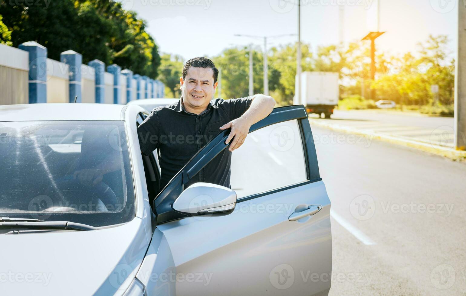 souriant chauffeur penché sur le voiture porte. content chauffeur penché sur le voiture porte dans le rue, souriant propriétaire avec Nouveau auto. Jeune Latin homme penché sur voiture porte dans le rue photo