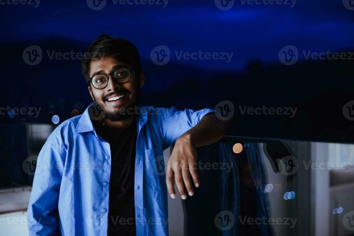 un Indien homme avec des lunettes et une bleu chemise regards autour le ville à nuit. dans le Contexte de le nuit rue de le ville photo