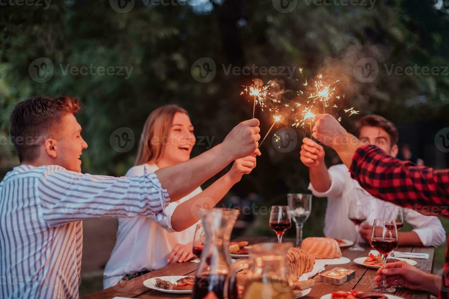 groupe de content copains célébrer vacances vacances en utilisant arroseurs et en buvant rouge du vin tandis que ayant pique-nique français dîner fête Extérieur près le rivière sur magnifique été soir dans la nature photo
