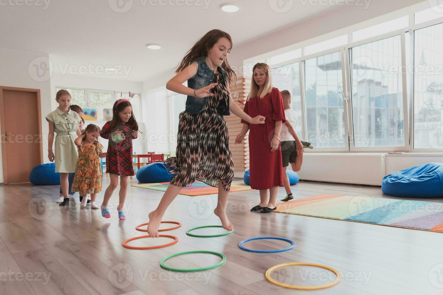 petit garderie école les enfants avec femelle prof sur sol à l'intérieur dans Salle de classe, Faire exercer. sauter plus de hula cerceau cercles Piste sur le sol. photo