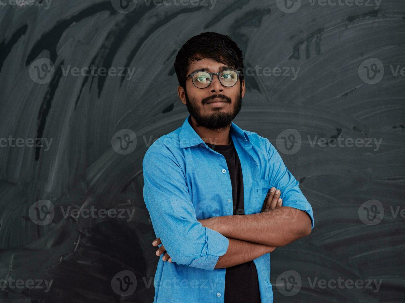 une Jeune Indien étudiant dans une bleu chemise avec des lunettes posant avec le sien bras franchi dans de face de le école tableau noir photo