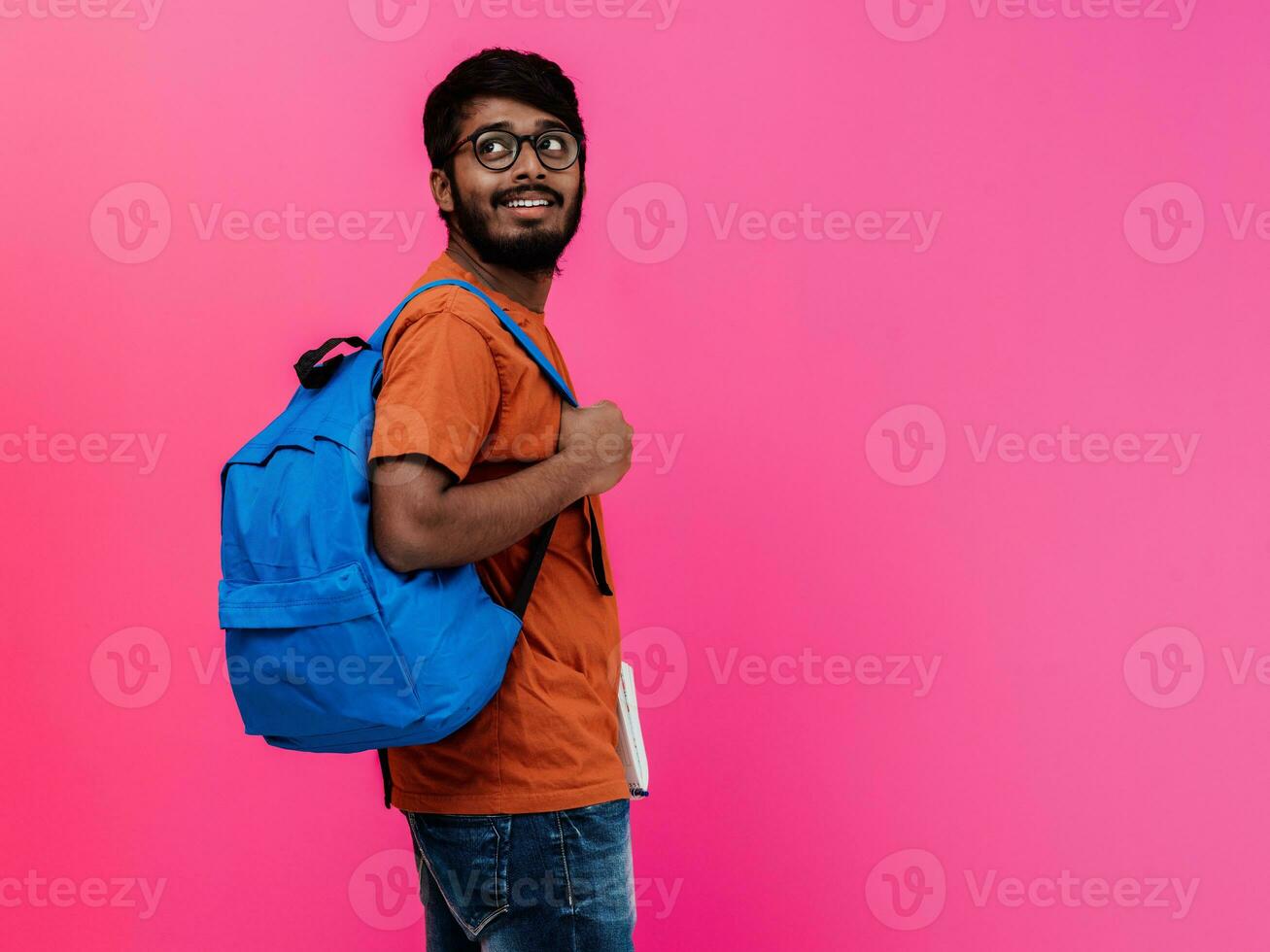 Indien étudiant avec bleu sac à dos, des lunettes et carnet posant sur rose Contexte. le concept de éducation et scolarité. temps à aller retour à école photo