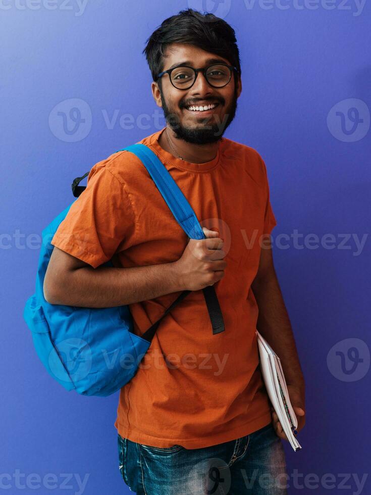 Indien étudiant avec bleu sac à dos, des lunettes et carnet posant sur violet Contexte. le concept de éducation et scolarité. temps à aller retour à école photo