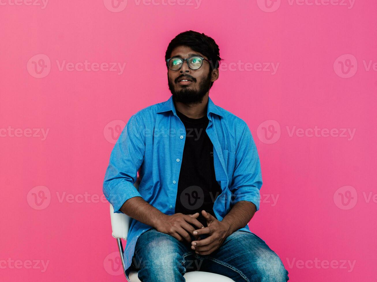 une Jeune Indien étudiant dans une bleu chemise avec des lunettes séance dans de face de une rose Contexte photo