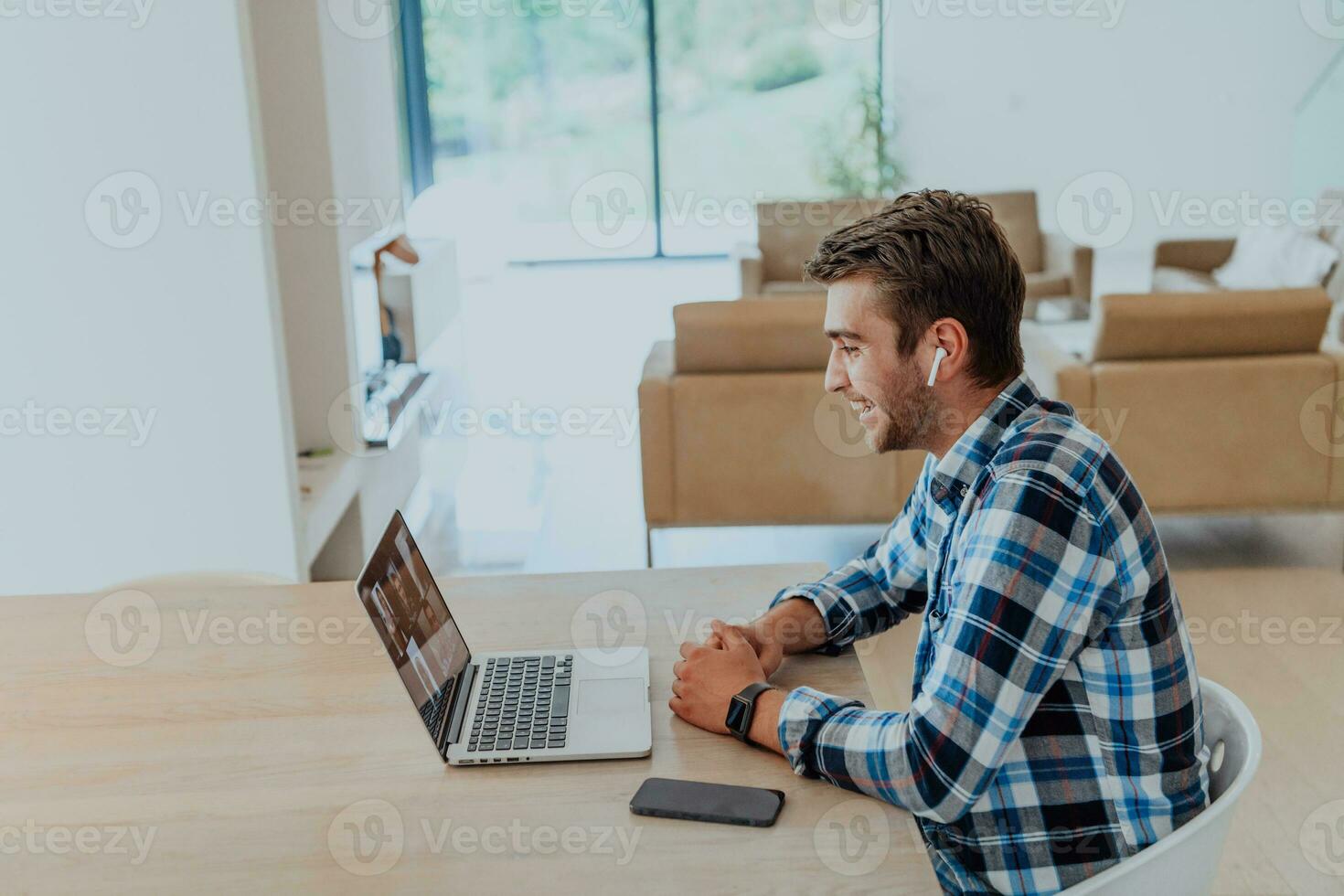 le homme séance à une table dans une moderne vivant chambre, avec écouteurs en utilisant une portable pour affaires vidéo discuter, conversation avec copains et divertissement photo