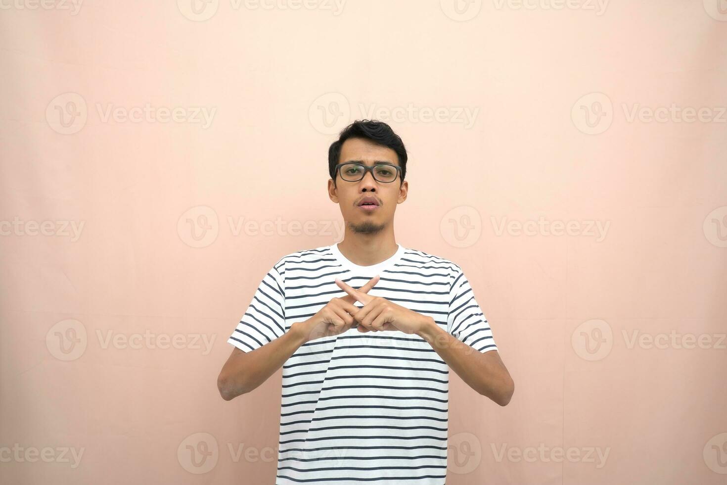 portrait de un asiatique homme dans des lunettes portant une décontractée rayé T-shirt. dénoter arrêt, traverser interdiction signe, interdire quelque chose, refuser,. beige Contexte. photo