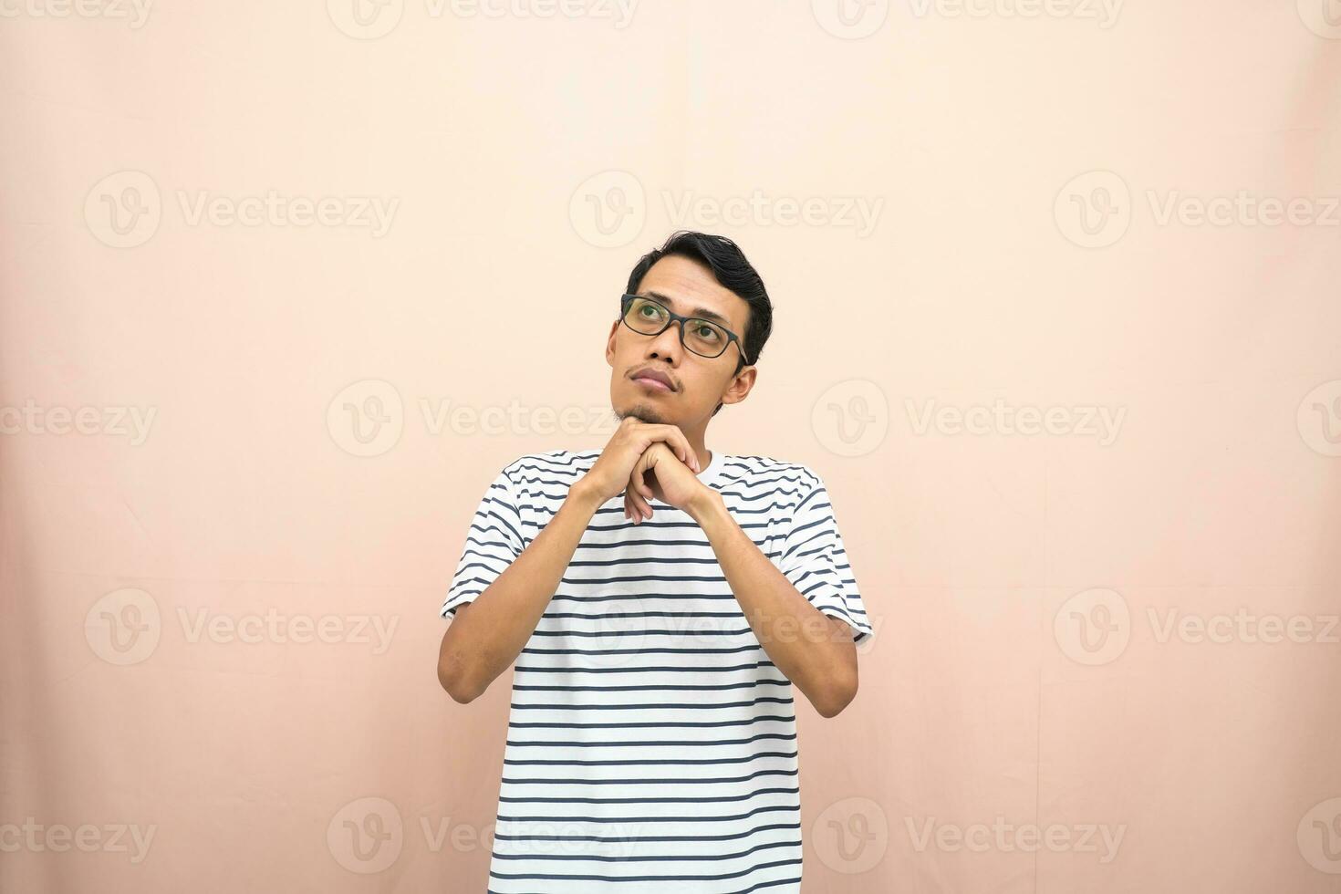 asiatique homme dans des lunettes portant décontractée rayé T-shirt, faire des gestes avoir des idées, à la recherche pour des idées, pensée. isolé beige Contexte. photo