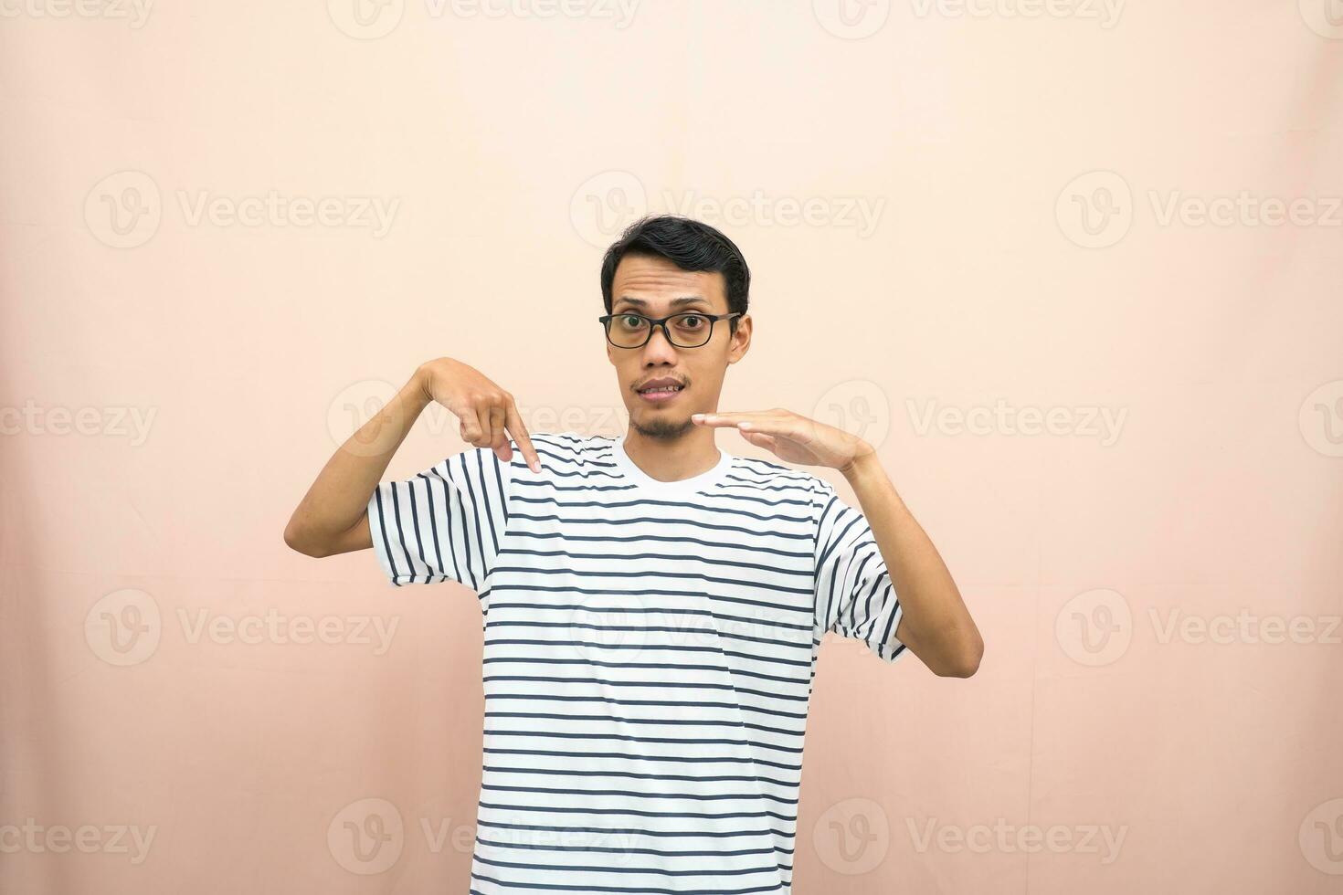 asiatique homme avec des lunettes portant décontractée rayé chemise, chuchotement pose tandis que montrer du doigt bas. isolé beige Contexte. photo