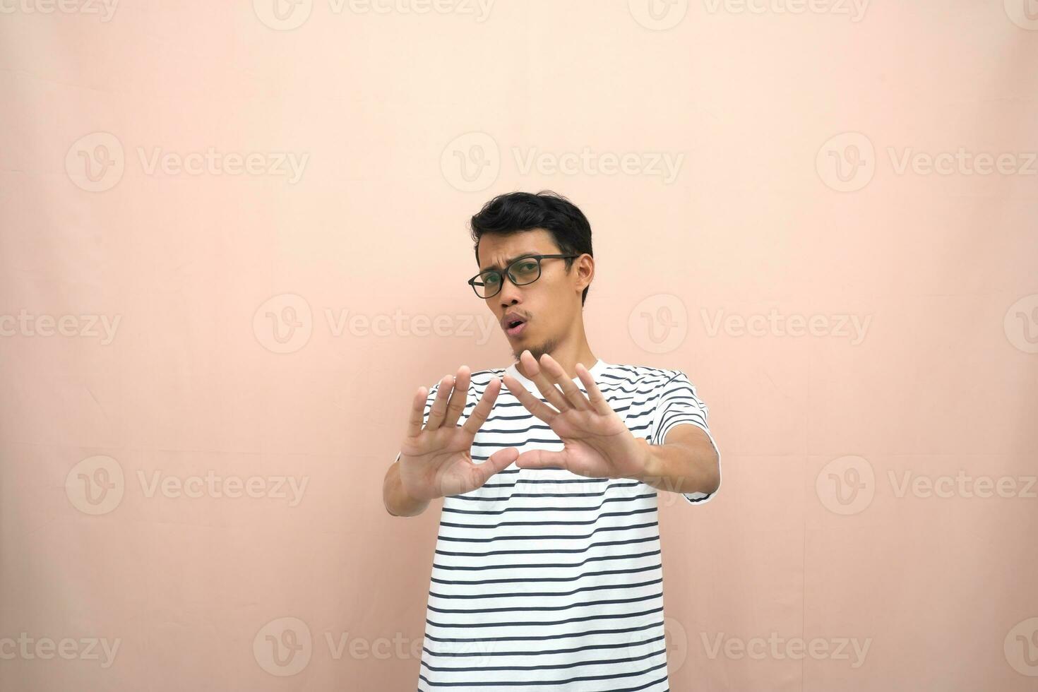 portrait de un asiatique homme dans des lunettes portant une décontractée rayé T-shirt. dénoter arrêt, traverser interdiction signe, interdire quelque chose, refuser,. beige Contexte. photo