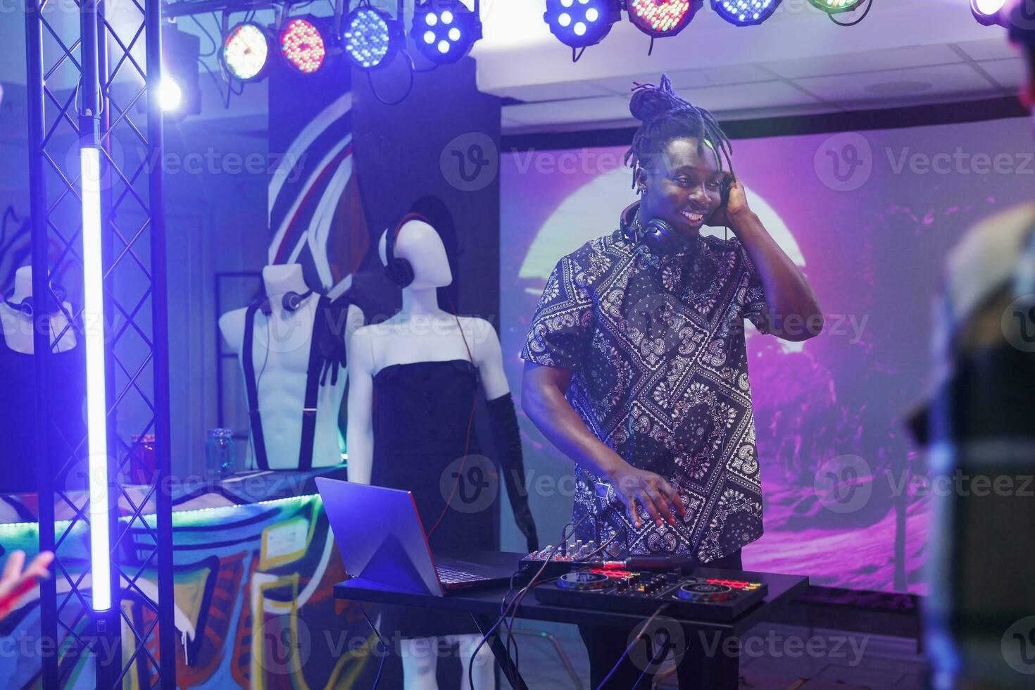 souriant musicien dans écouteurs en jouant sur boîte de nuit étape avec projecteurs. africain américain homme dj performant en utilisant électronique la musique manette à discothèque fête dans club photo