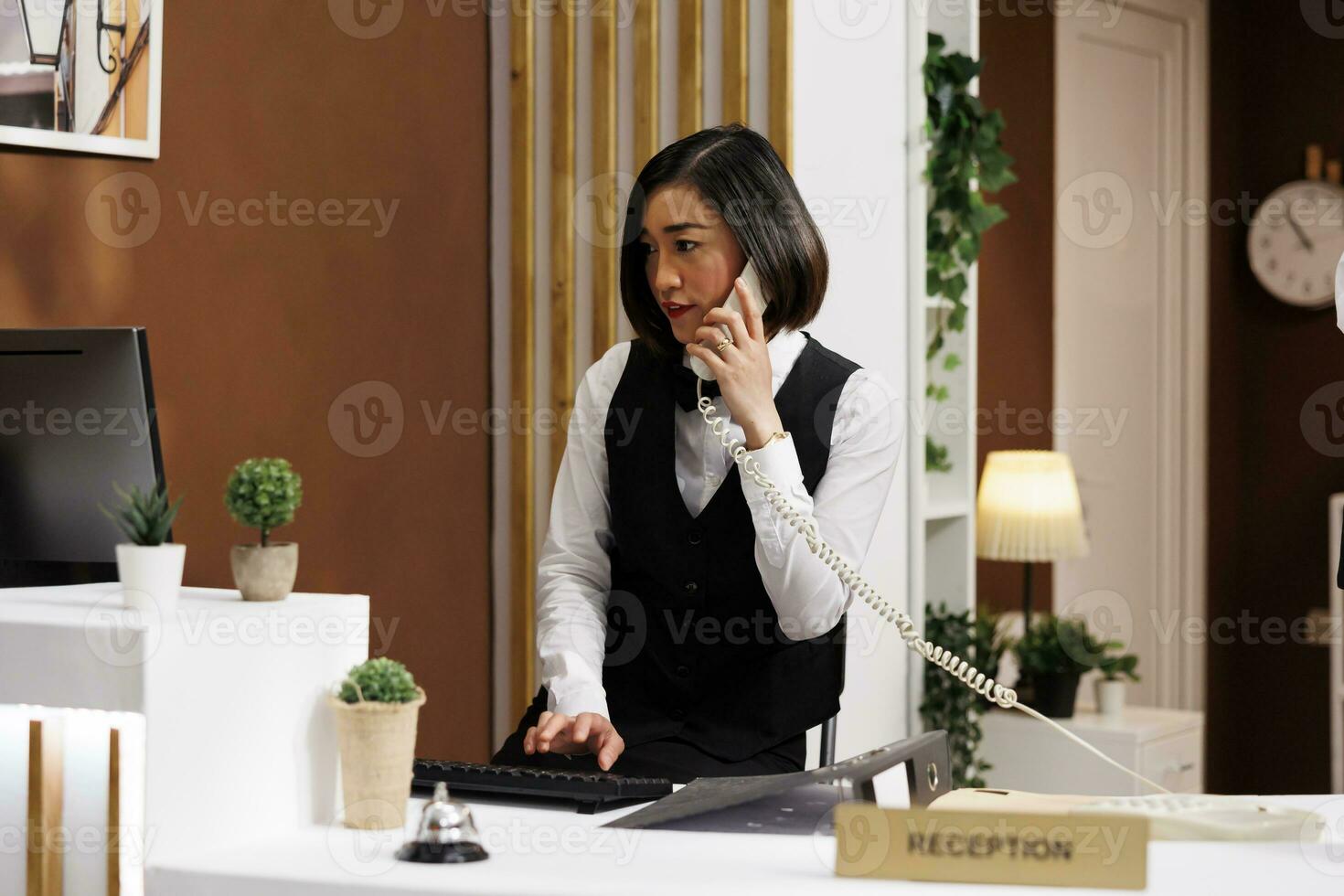 femme réceptionniste habillé dans uniforme, parlant sur le téléphone à de face bureau, assistant les clients avec réservation chambre. Jeune asiatique la conciergerie ayant téléphone conversation avec client photo