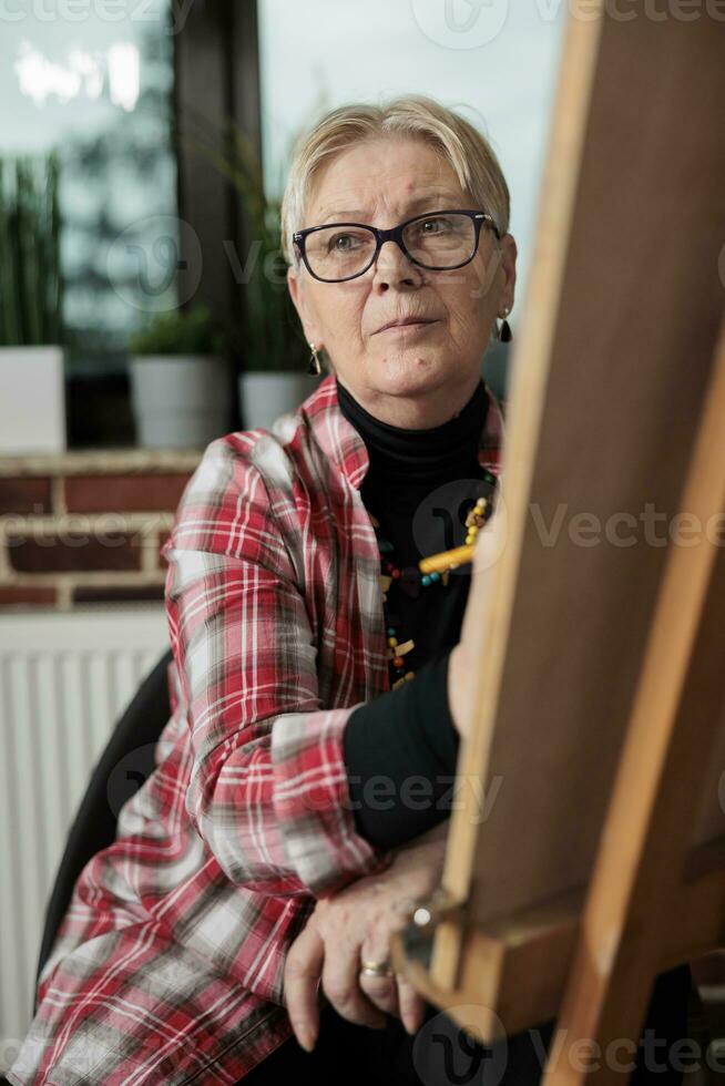 art Des classes pour personnes âgées. pensif mature femme séance dans de face de chevalet à la recherche de côté, écoute à prof pendant dessin leçon, apprentissage esquisse dans petit groupes. plus âgée gens et la créativité photo