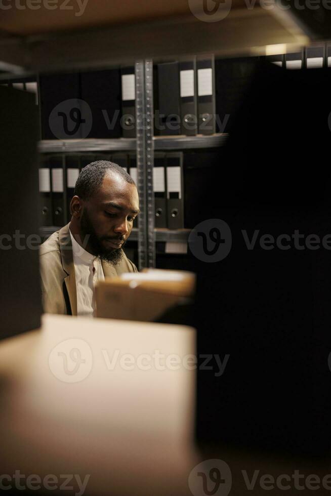 loi mise en vigueur professionnel travail dans foncé Bureau en retard et séance à lieu de travail plein de étagères avec Dossiers. concentré africain américain police détective résolution la criminalité à nuit temps photo