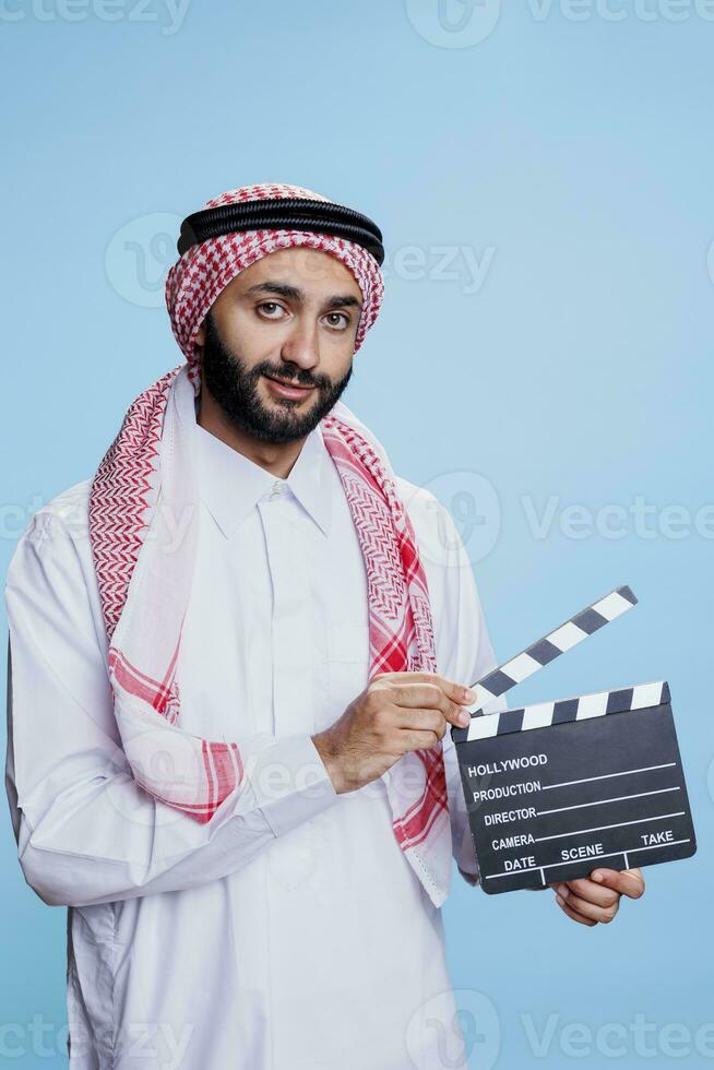 musulman homme dans traditionnel vêtements posant avec film ardoise et à la recherche à caméra avec souriant expression. arabe la personne mettant en valeur film production clap studio portrait photo