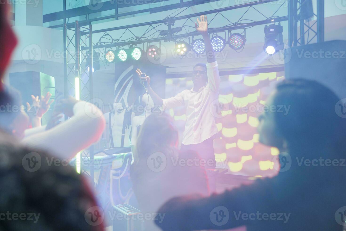 musicien avec microphone élevage main tandis que divertissant foule de étape dans boîte de nuit. dj en chantant et en jouant à électronique la musique spectacle et discothèque fête dans club avec projecteurs photo