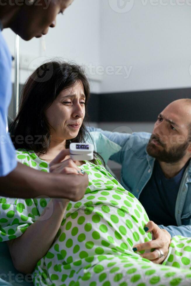 Enceinte femme ayant douloureux contractions étant aidé et réconfortant par africain américain infirmière et mari dans hôpital salle. patient avec grossesse mensonge dans lit en train de préparer à donner naissance photo