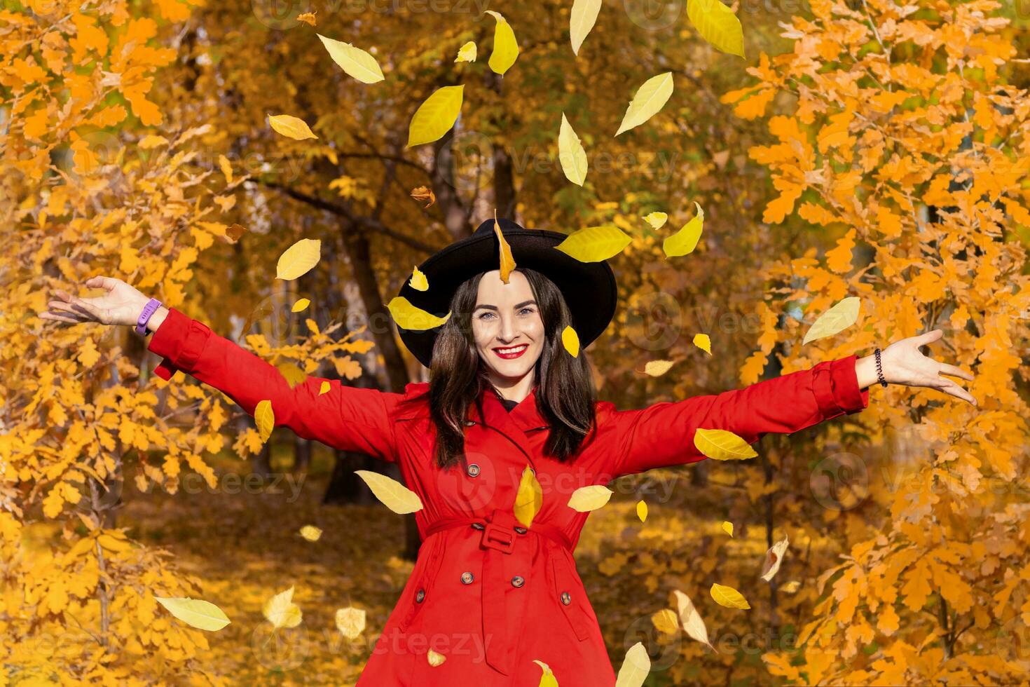 souriant femme dans une noir chapeau et avec rouge rouge à lèvres lancement Jaune feuilles en haut dans le l'automne forêt parc photo