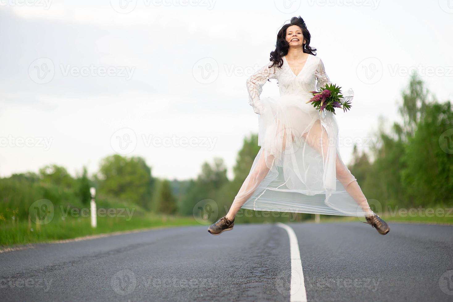 drôle de jeune femme en robe blanche sautant sur une route goudronnée vide photo