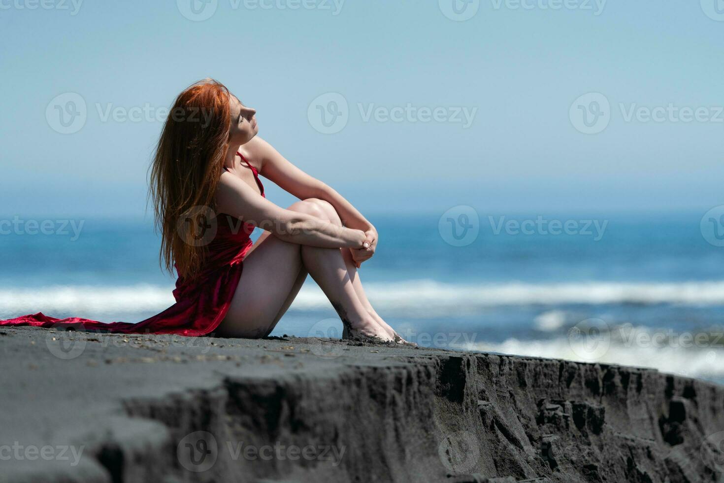 femme dans rouge longue robe avec élevé ourlet et nu jambes séance sur noir le sable sur plage, étreindre les genoux photo