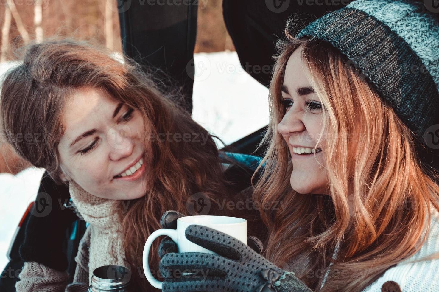 amis heureux dans la forêt d'hiver. deux filles s'asseyent dans le tronc buvant du café photo