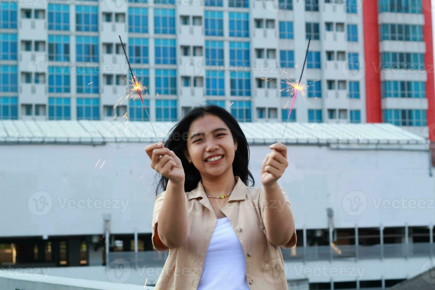 insouciant asiatique Jeune femme en portant cierge magique célébrer Nouveau année veille avec rire et Danse dans toit appartement Extérieur avec ville bâtiment Contexte photo