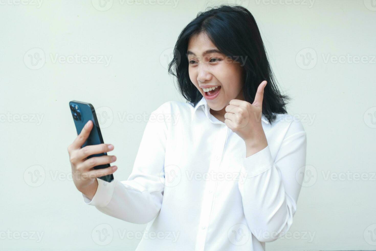 portrait de excité Jeune asiatique femme entrepreneur donnant les pouces en haut souriant espiègle satisfait à la recherche à téléphone intelligent détient sur mains portant blanc costume chemise permanent isolé, bien un service geste photo