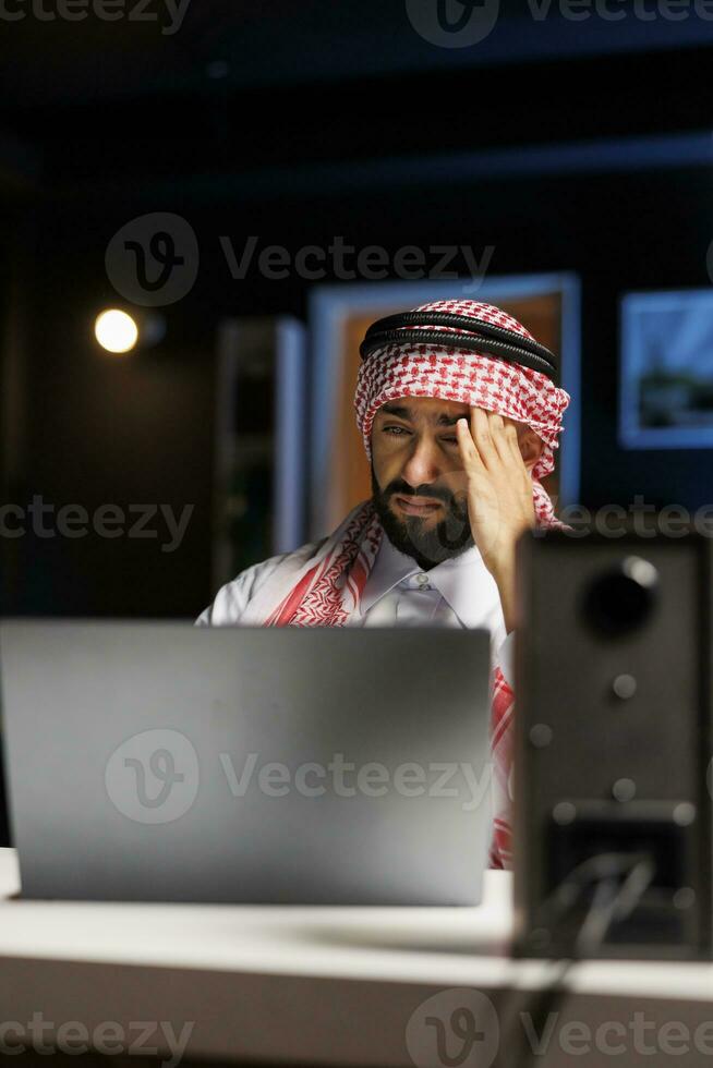 détaillé vue de fatigué homme dans arabe tenue séance à Bureau bureau et en utilisant portable à conduite l'Internet recherche. musulman homme avec mini-ordinateur sur le tableau, à la recherche se lasser et Souffrance de mal de tête. photo