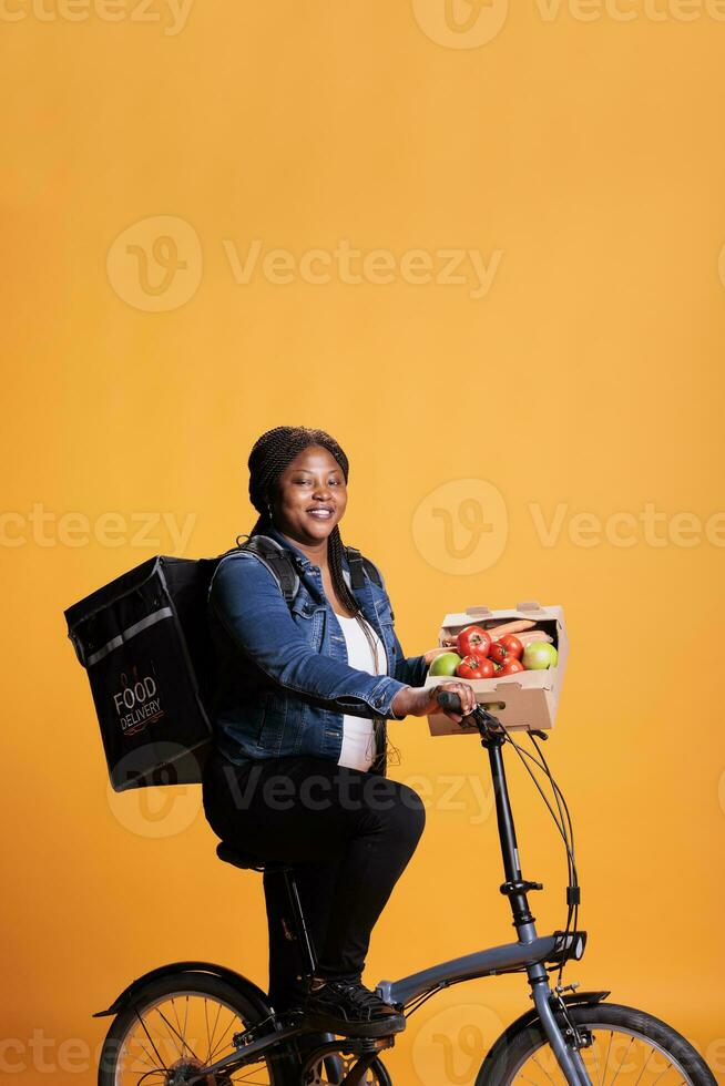 africain américain épicerie livreuse équitation bicyclette tandis que livrer boîte plein avec biologique des légumes à client pour en bonne santé le déjeuner tandis que en utilisant bicyclette comme transport. prendre en dehors nourriture un service et concept photo