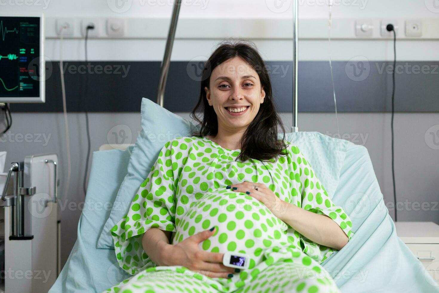 souriant Enceinte femme mensonge dans lit en portant mains sur ventre dans hôpital salle. de bonne humeur patient avec ventre grossesse à la recherche à caméra et attendre sur médecin à faire examen pendant vérification visite photo