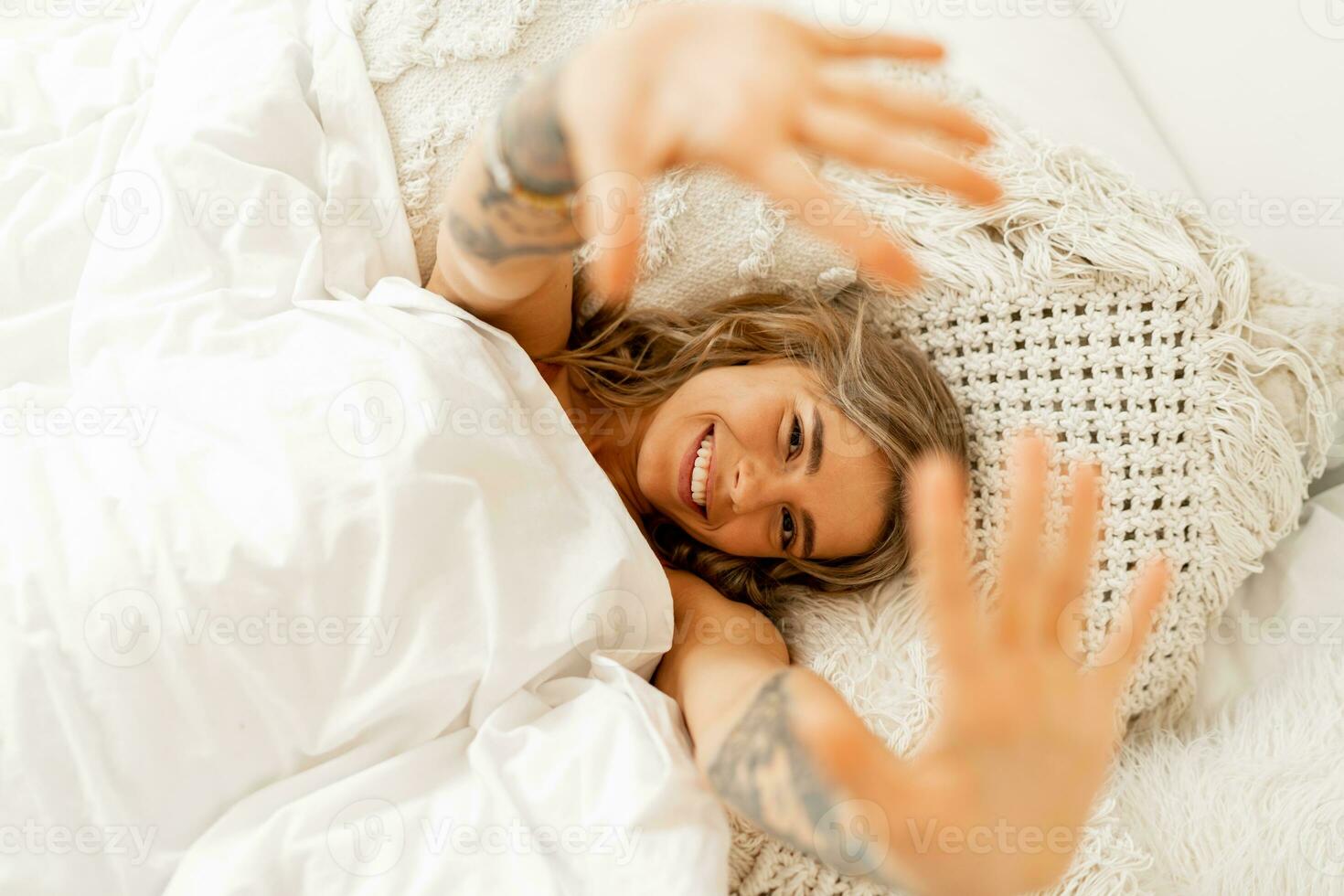 Haut vue portrait de content de bonne humeur femme mensonge dans blanc confortable lit réveiller en haut dans le matin, tire mains avant. chaud couleurs, boho intérieur. photo